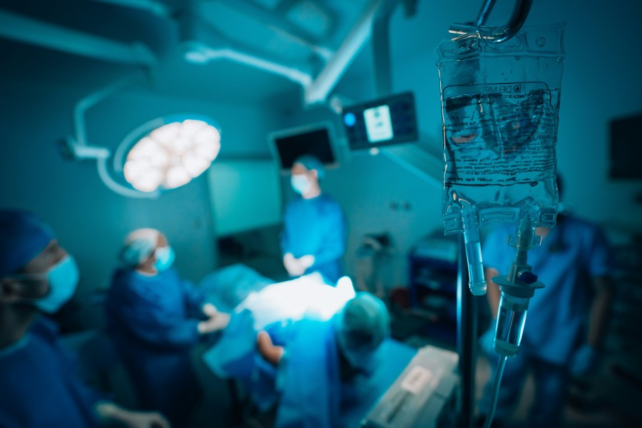 Surgical team performing an operation in a brightly lit operating room, with an IV fluid bag in the foreground.