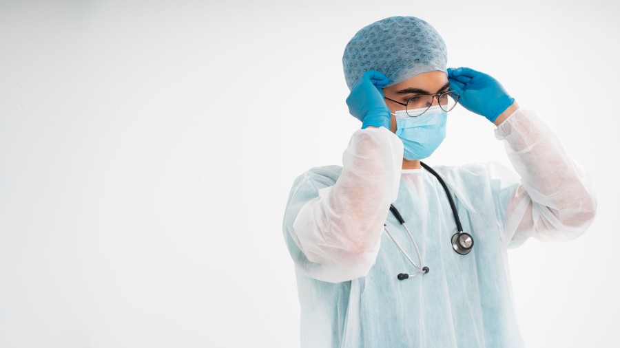 Healthcare professional in protective medical gown, mask, gloves, and hair cover adjusting protective eyewear.