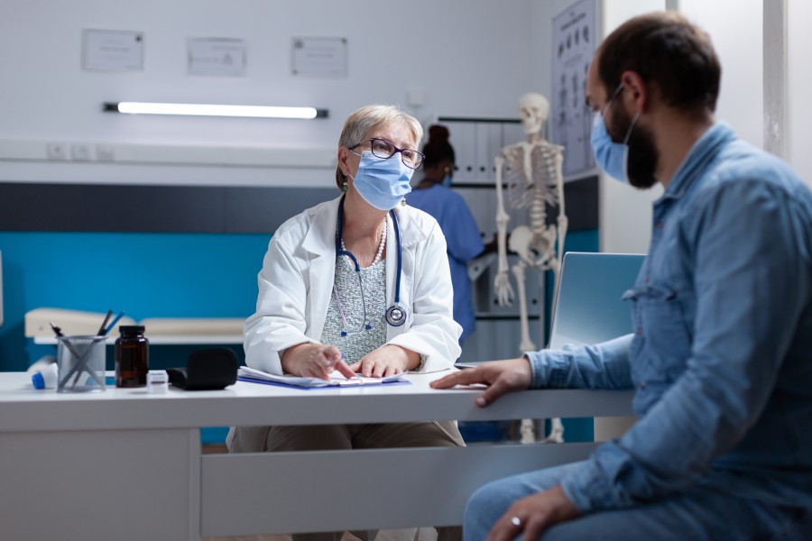 Doctor wearing a face mask consulting with a masked male patient in a medical examination room.